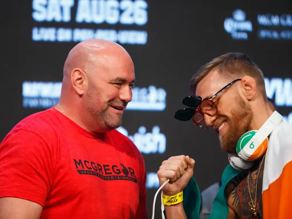 Two men fist-bump on a backstage stage; one in a red McGregor shirt smiles, the other wearing sunglasses and headphones draped with an flag.