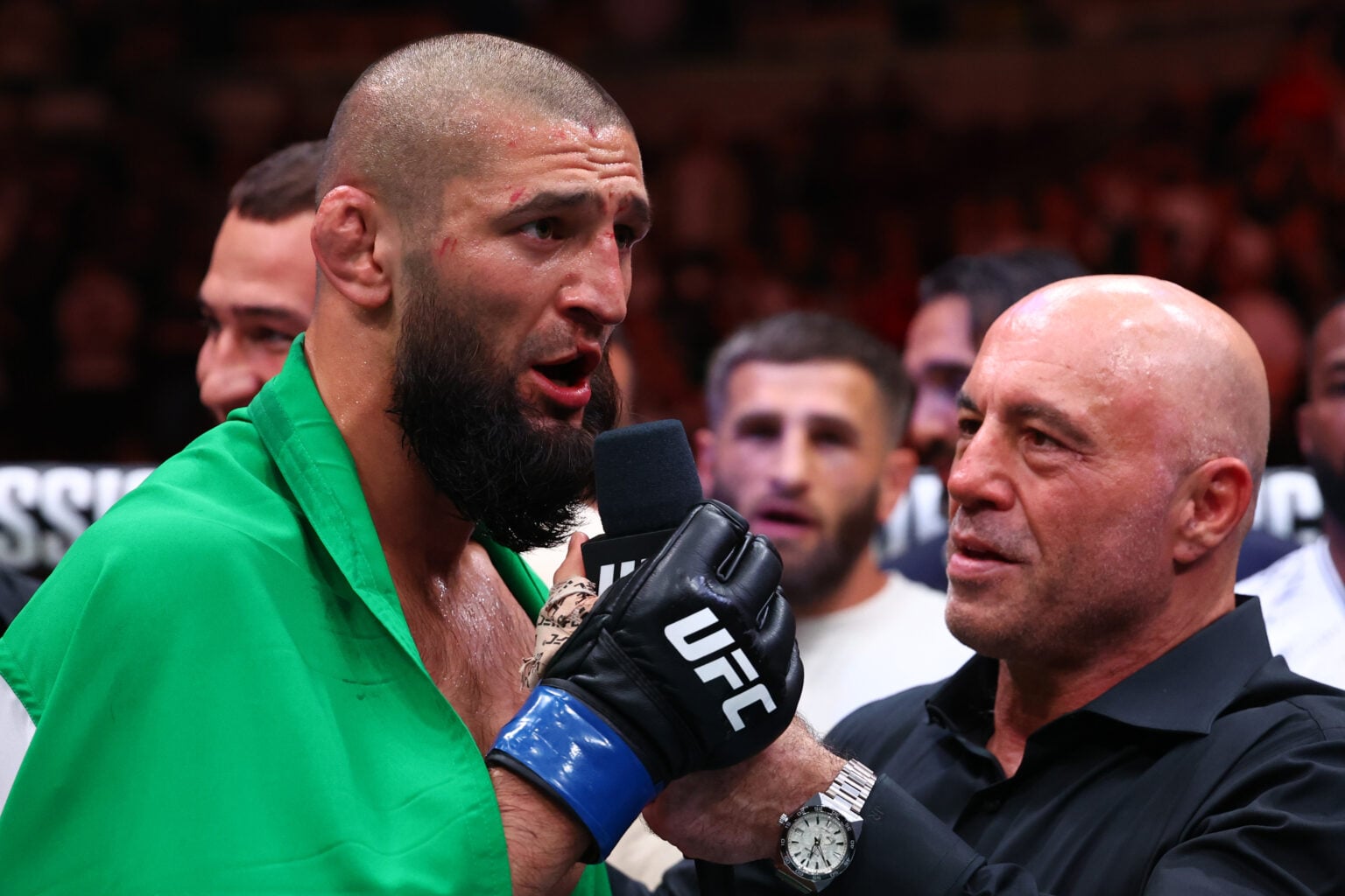 Bearded fighter in a green robe speaks into a microphone held by an interviewer in a black shirt; crowd in the background.