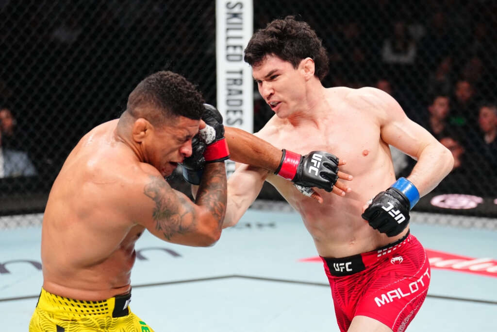 Two shirtless UFC fighters exchange a punch inside the octagon, one in red shorts blocking with a forearm while the other in yellow shorts recoils.