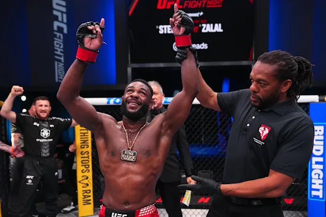 Shirtless UFC fighter with red gloves celebrates victory with arms raised inside the cage, a referee stands nearby, and teammates cheer in the background.