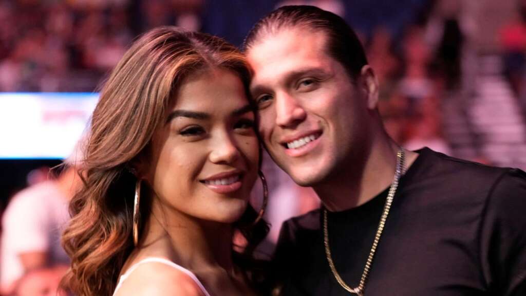 Smiling couple posing closely for a photo at a crowded venue; woman with long hair and large hoop earrings, man beside her wearing a chain.