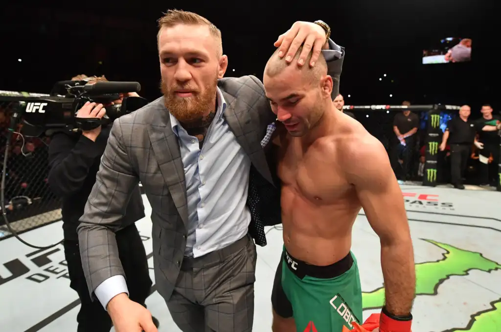 624421900 - AfroCombat Bearded man in a gray suit supports a shirtless fighter with green shorts inside the UFC ring after a bout.
