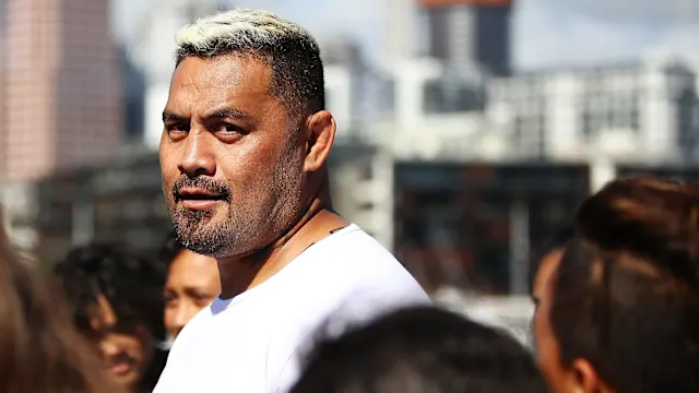 Man with short gray-blonde hair and a white shirt stands among a crowd outdoors in an urban area.