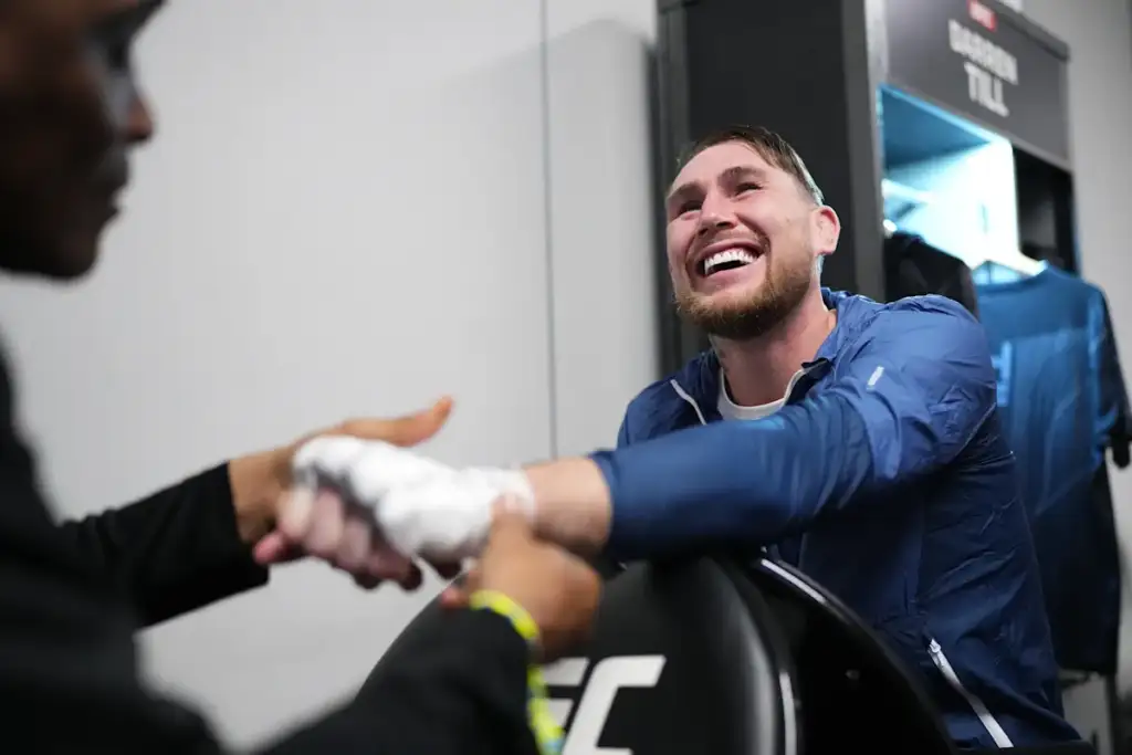 Athlete in a blue jacket smiles as he shakes hands with a trainer in a locker-room gym.