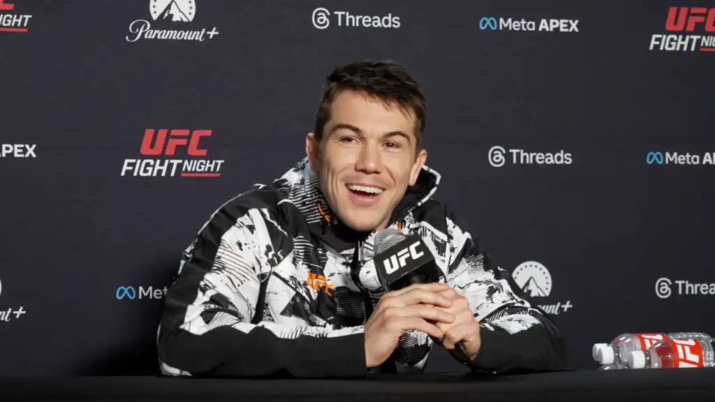 UFC fighter smiles at a press conference, holding a microphone, wearing a black-and-white patterned jacket with sponsor logos on the backdrop behind him.