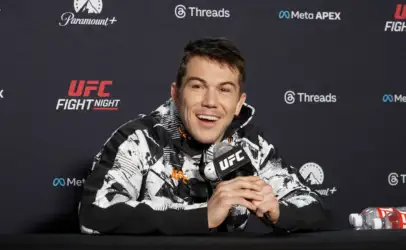 UFC fighter smiles at a press conference, holding a microphone, wearing a black-and-white patterned jacket with sponsor logos on the backdrop behind him.