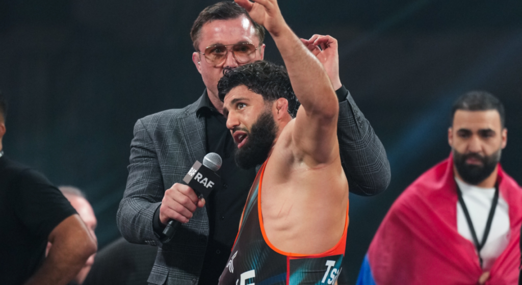 Wrestler with orange-trim singlet raises arms in victory while interviewer holds a mic labeled RAF nearby.
