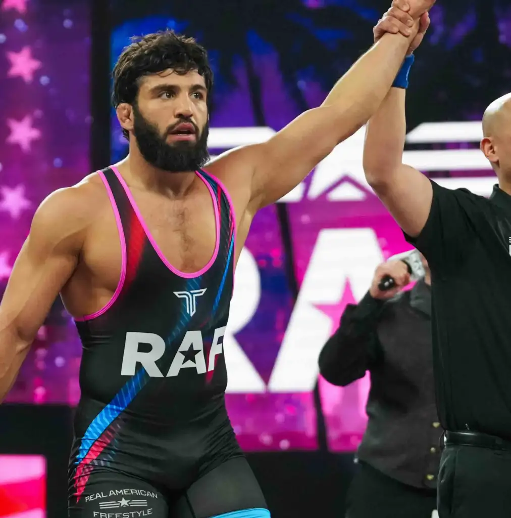 Bearded male wrestler in a black-pink singlet with 'Real American Freestyle' on the shorts, arms raised in victory with a teammate/referee nearby.
