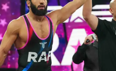 Bearded male wrestler in a black-pink singlet with 'Real American Freestyle' on the shorts, arms raised in victory with a teammate/referee nearby.