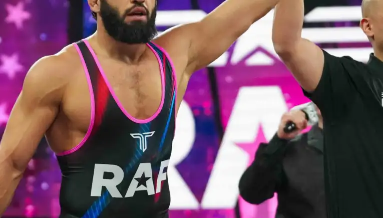 Bearded male wrestler in a black-pink singlet with 'Real American Freestyle' on the shorts, arms raised in victory with a teammate/referee nearby.