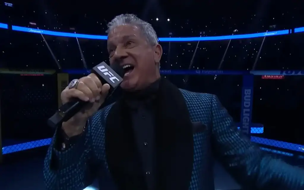 UFC announcer speaking into a UFC-branded microphone inside the octagon, wearing a blue patterned blazer.