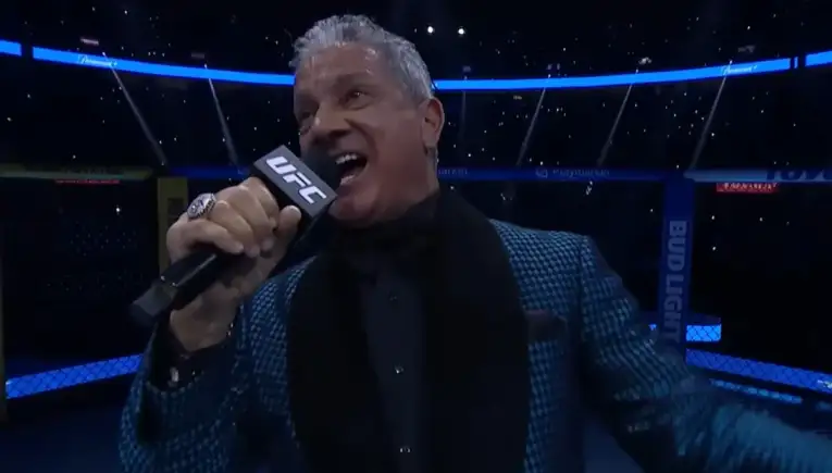 UFC announcer speaking into a UFC-branded microphone inside the octagon, wearing a blue patterned blazer.