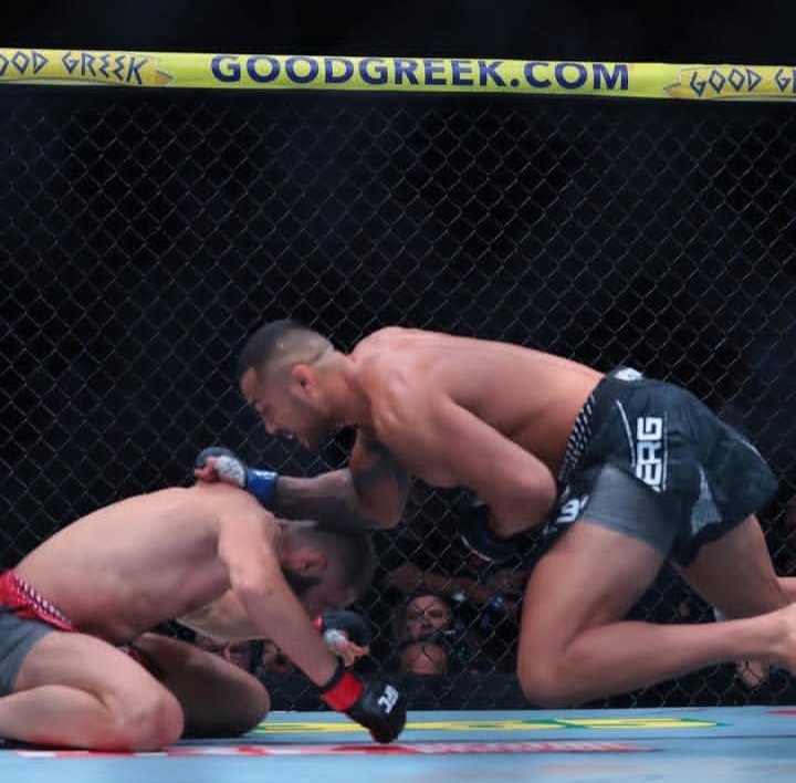 Two male fighters in a cage MMA match; one on the mat shielding head as the other punches in.