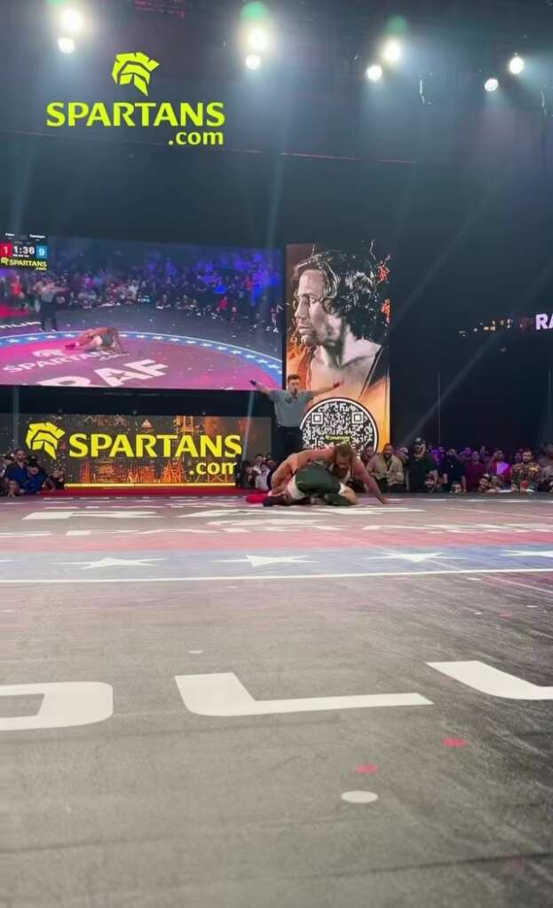 Wrestler on a mat during a Spartan event, with SPARTANS signage and a large crowd in the arena.