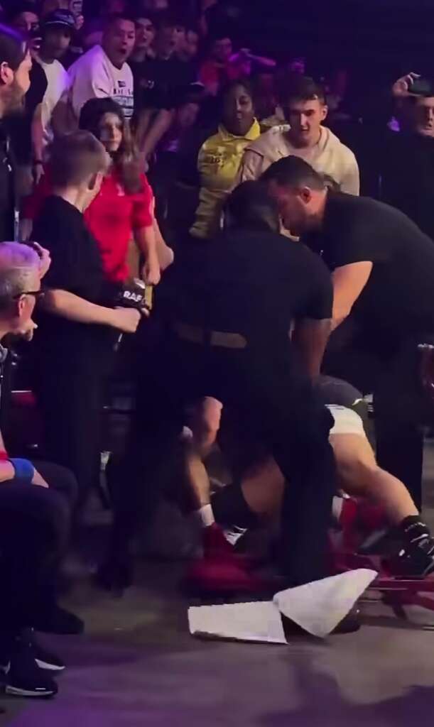 Crowd in a dim arena watches as two men in black assist a person on the floor in white shorts, with a red mat nearby and scattered papers on the ground.