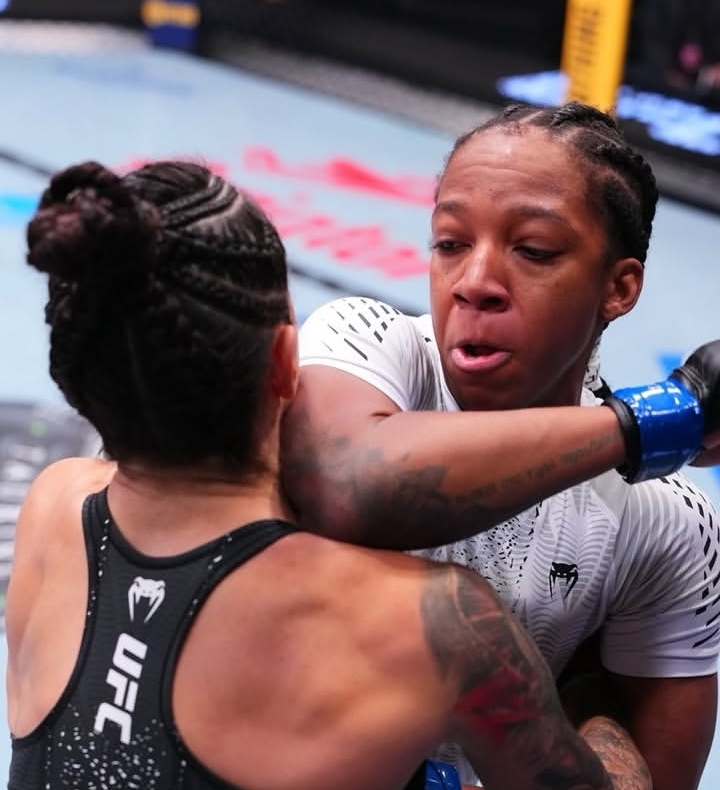 Two female UFC fighters grapple in the octagon during a match; one in white with braided hair, the other in black, with a blue-gloved arm near them.