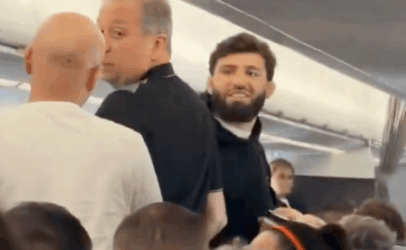 Crowded airplane aisle with a bearded man in a dark shirt smiling at the camera amid surrounding passengers.