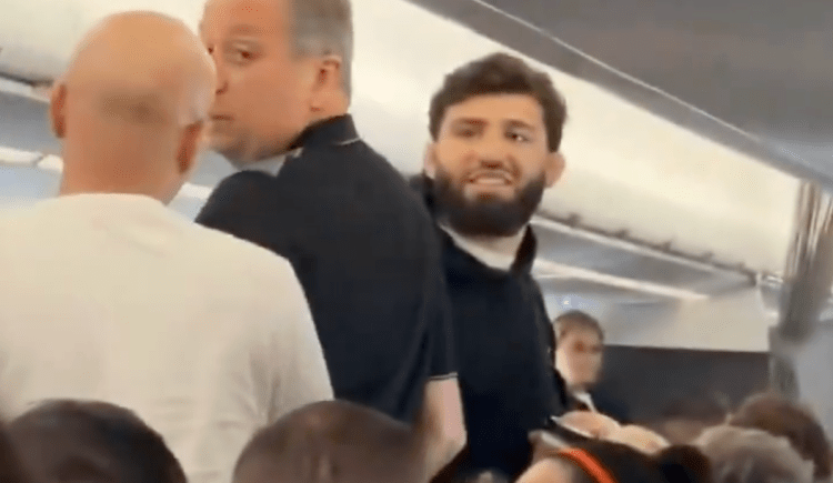 Crowded airplane aisle with a bearded man in a dark shirt smiling at the camera amid surrounding passengers.