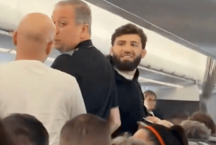 Crowded airplane aisle with a bearded man in a dark shirt smiling at the camera amid surrounding passengers.