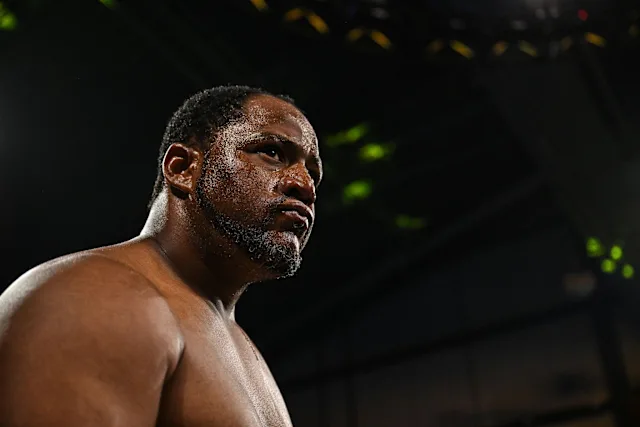 Close-up of a muscular male boxer with a beard, looking focused in a dim arena under lights.