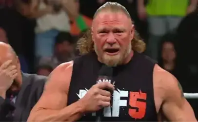 Muscular wrestler in a black sleeveless shirt holding a microphone and shouting in a wrestling ring.