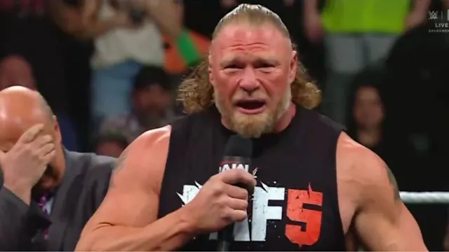 Muscular wrestler in a black sleeveless shirt holding a microphone and shouting in a wrestling ring.