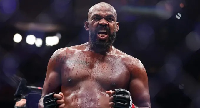 Intense shirtless male fighter with fists clenched, shouting in a ring under bright arena lights.