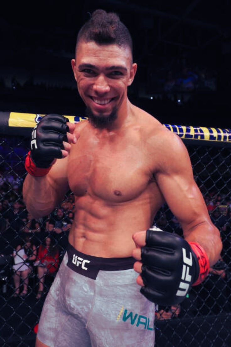 UFC fighter posing shirtless in the octagon, fists up and smiling at the camera.