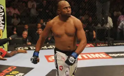 Male mixed martial artist standing in the cage, wearing blue gloves and white shorts with logos, looking toward the crowd between rounds.