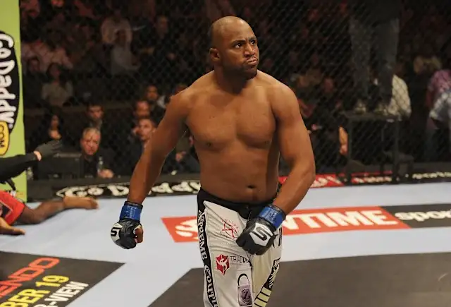 Male mixed martial artist standing in the cage, wearing blue gloves and white shorts with logos, looking toward the crowd between rounds.