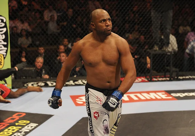 Male mixed martial artist standing in the cage, wearing blue gloves and white shorts with logos, looking toward the crowd between rounds.