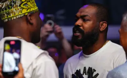 Close-up of two men in conversation at a crowded event; right man has a beard and earring, left man wears a yellow patterned headwrap, foreground phone visible