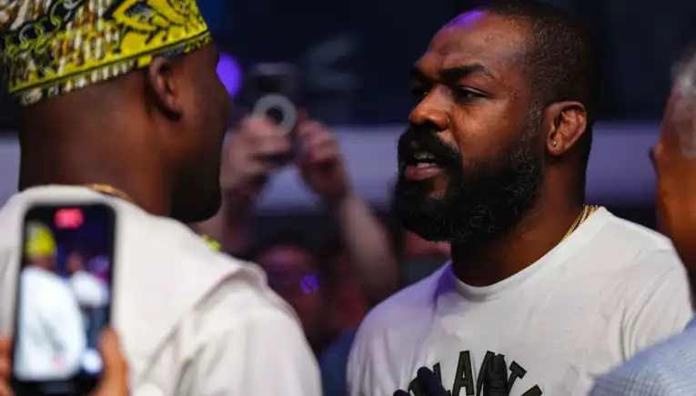 Close-up of two men in conversation at a crowded event; right man has a beard and earring, left man wears a yellow patterned headwrap, foreground phone visible