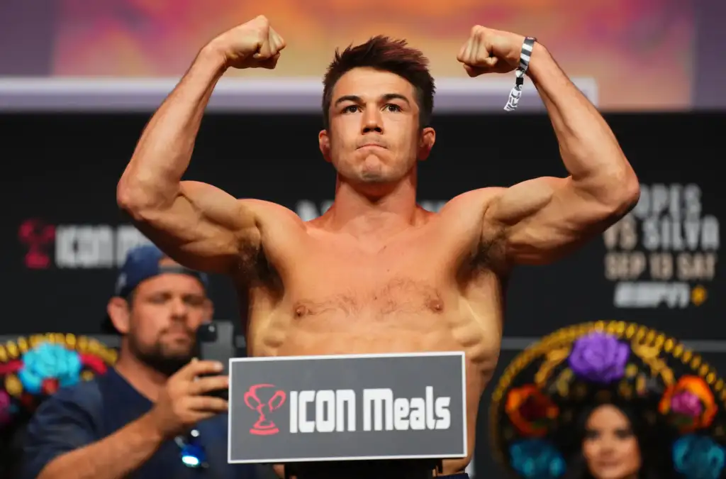 Shirtless male bodybuilder on stage flexing arms above his head, holding a sign that reads ICON Meats in the foreground.