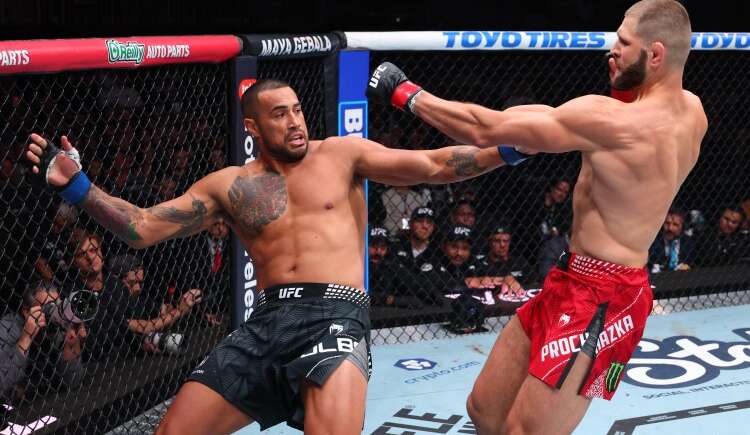 Two UFC fighters exchange punches inside the cage during a match: left fighter in black shorts with tattoos, right fighter in red shorts lunging with a punch.