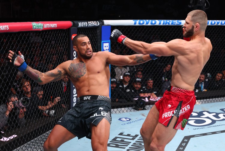 Two UFC fighters exchange punches inside the cage during a match: left fighter in black shorts with tattoos, right fighter in red shorts lunging with a punch.