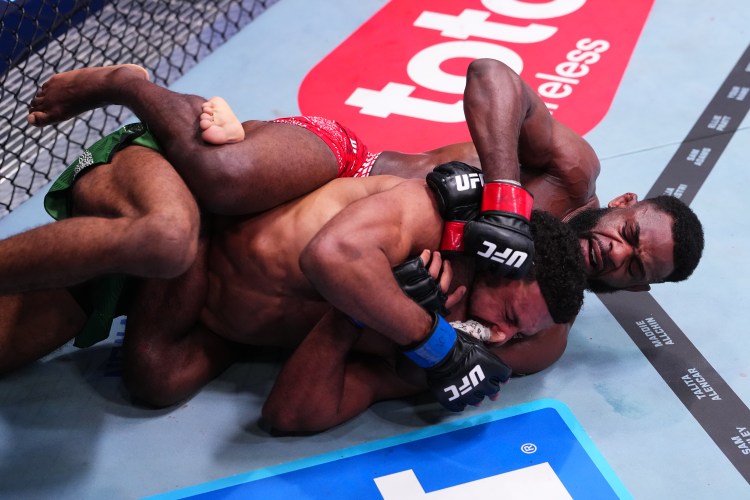 Two UFC fighters grapple on the mat during a match, one in red shorts on top and the other in green shorts beneath, inside the cage.