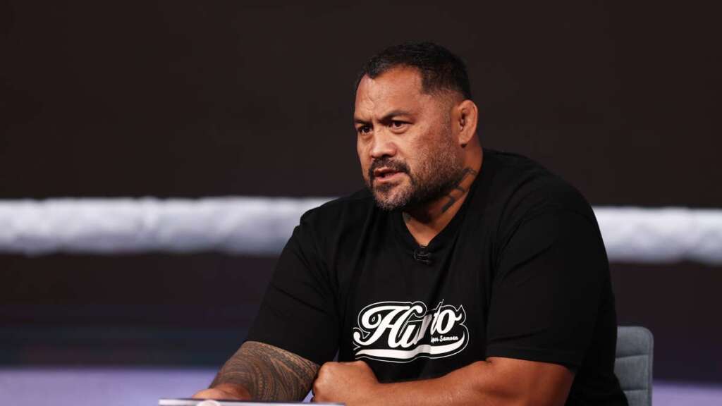 Man with short hair and a beard, wearing a black T-shirt, speaks during an interview in a studio setting