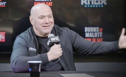 Man in a black jacket speaks into a UFC microphone at a press table, with a dark backdrop and coffee cup nearby.