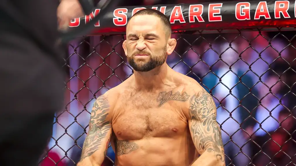 MMA fighter inside a chain-link cage, shirtless, eyes squeezed shut and grimacing, tattoos visible on arms and chest
