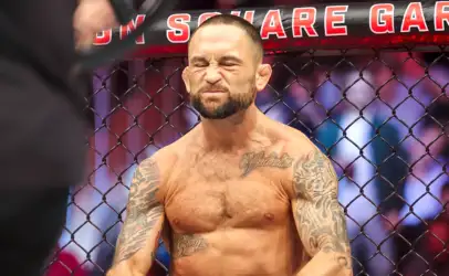 MMA fighter inside a chain-link cage, shirtless, eyes squeezed shut and grimacing, tattoos visible on arms and chest
