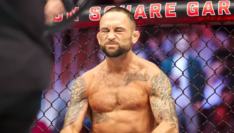 MMA fighter inside a chain-link cage, shirtless, eyes squeezed shut and grimacing, tattoos visible on arms and chest
