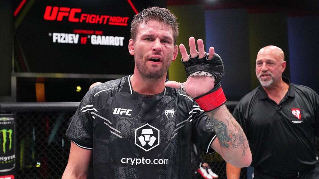 UFC fighter wearing a crypto.com shirt raises his hand in victory inside the cage, with a coach in the background.