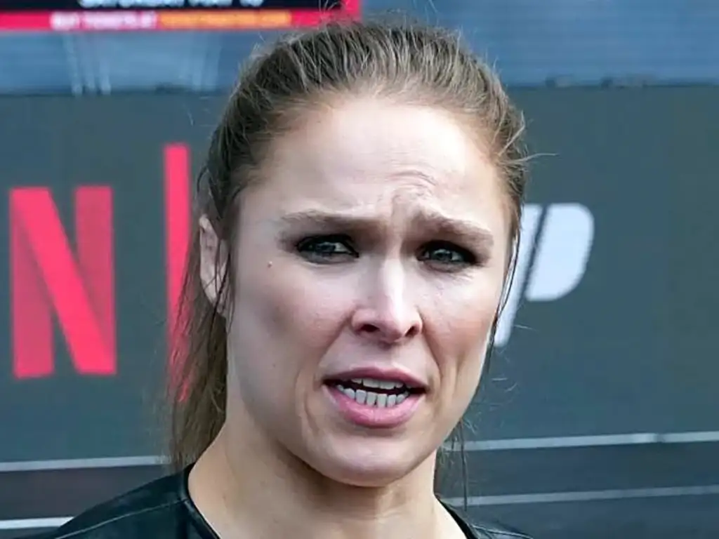 Close-up of a woman with a tense expression speaking outdoors, wearing a dark top.