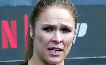 Close-up of a woman with a tense expression speaking outdoors, wearing a dark top.