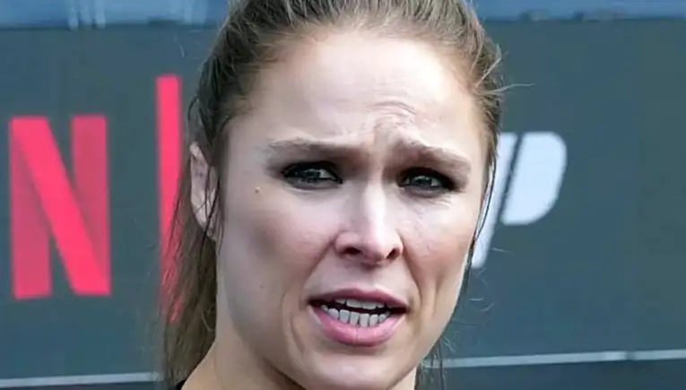 Close-up of a woman with a tense expression speaking outdoors, wearing a dark top.