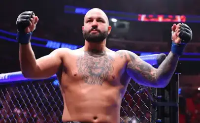 Professional mixed martial arts fighter with tattoos and gloves, shirtless, standing inside the octagon with arms raised in victory.