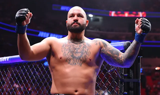 Professional mixed martial arts fighter with tattoos and gloves, shirtless, standing inside the octagon with arms raised in victory.