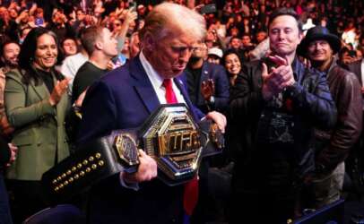Donald Trump in a suit and red tie holds a wrestling championship belt as a crowd cheers around him.