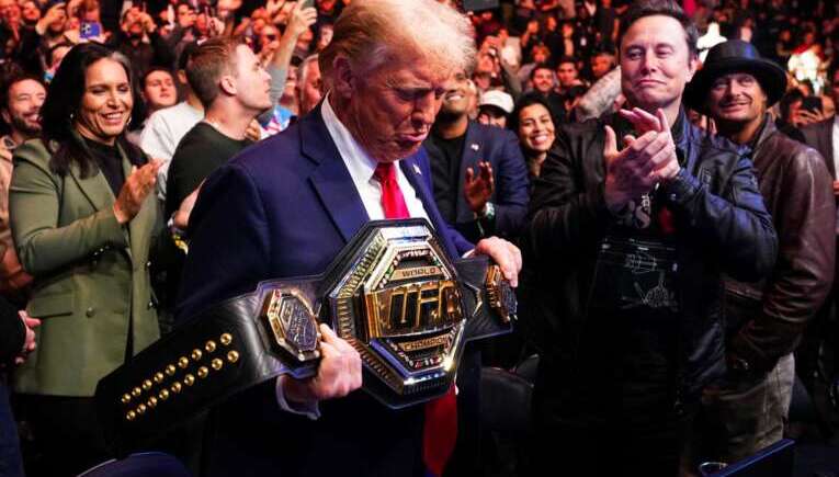 Donald Trump in a suit and red tie holds a wrestling championship belt as a crowd cheers around him.